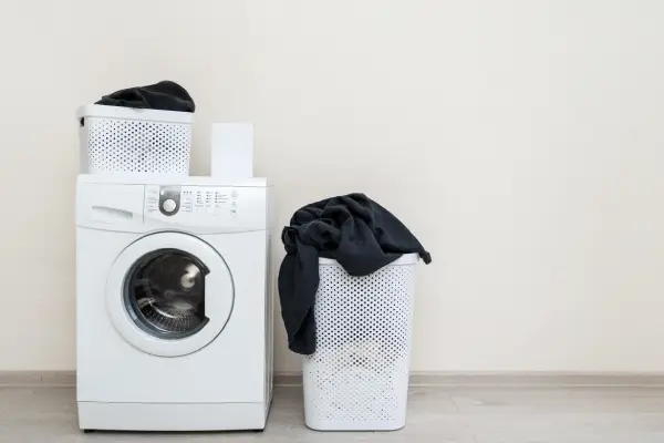 Preparation to laundry process. White washing machine with box of detergent soap and black clothes in basket standing on floor inside bright light flat interior